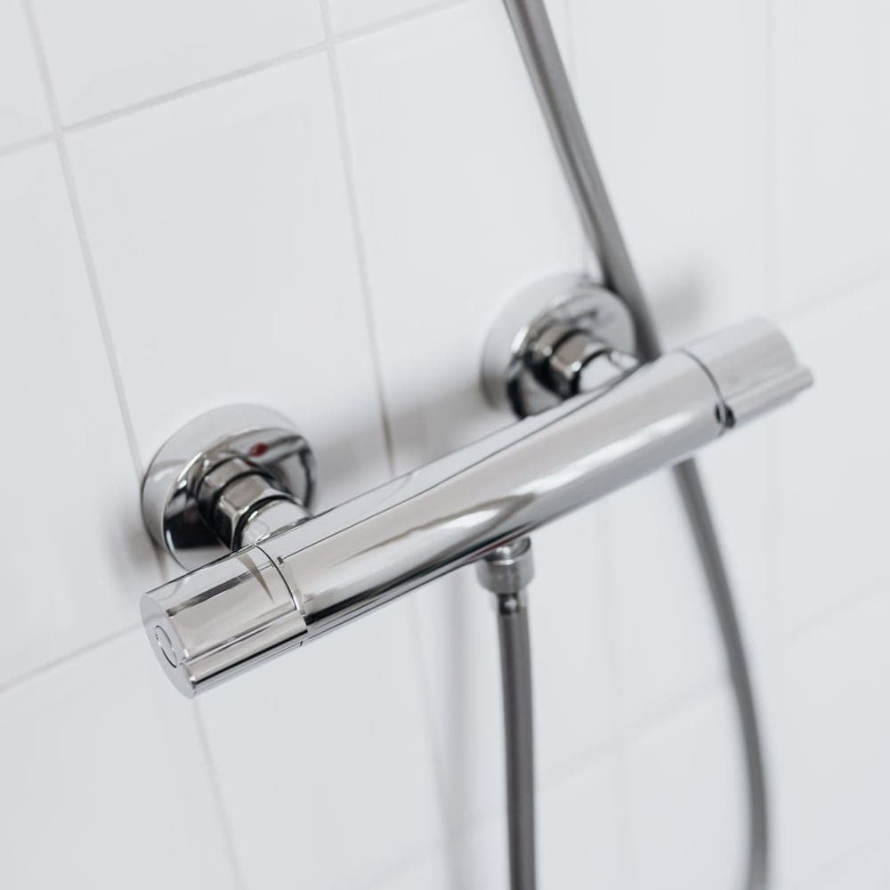 Grifo termostático de ducha cromado sobre azulejo blanco, con el flexible colgando
